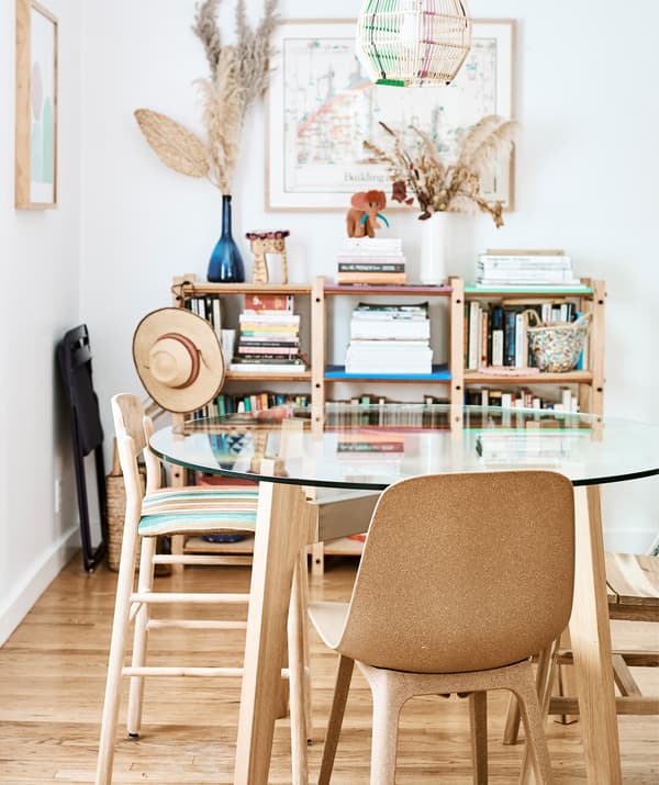 Coin repas avec une table à plateau de verre et des chaises brunes, et des étagères contre un mur blanc.