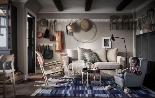 Coin salon avec canapé, fauteuil à bascule et un garçon dans un fauteuil d’enfant. Sur le mur, alignement de patères pour rangement/exposition.