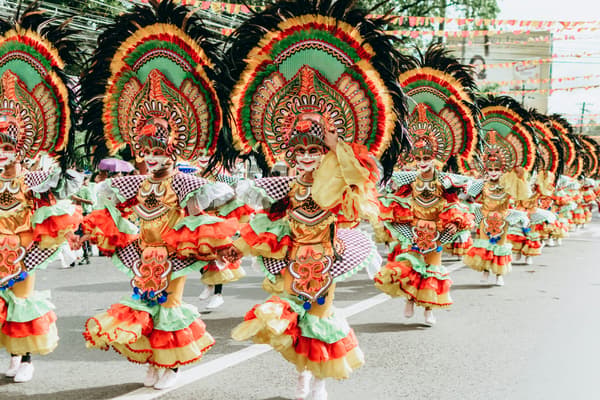 Colorfully dressed performers in vibrant costumes and elaborate headdresses dance in a festive parade along a city street.