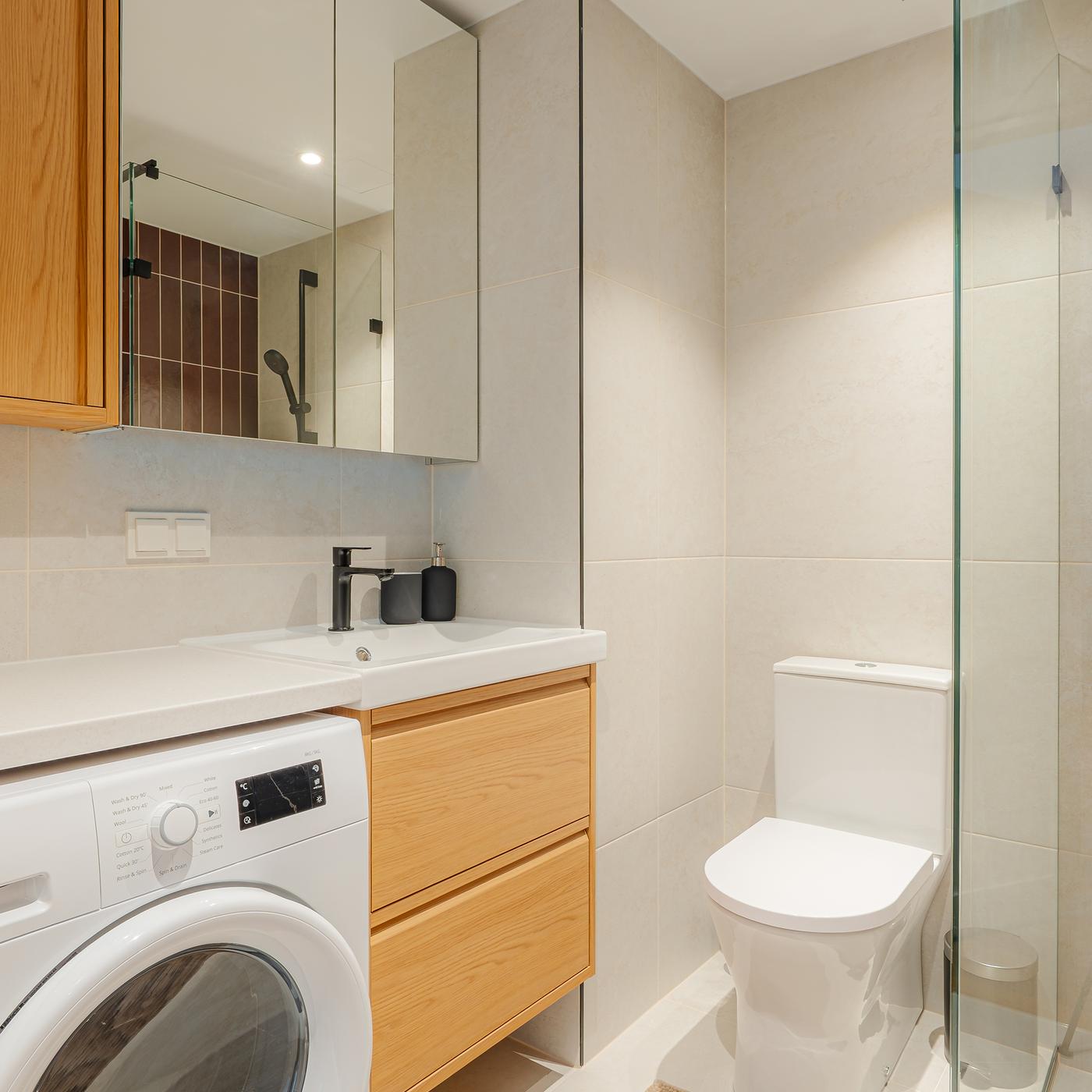Compact bathroom with an oak effect wash stand and wall cabinet, black tap and shower fittings, mirrored cabinets, a washing machine below a neutral countertop, and a glass shower enclosure.