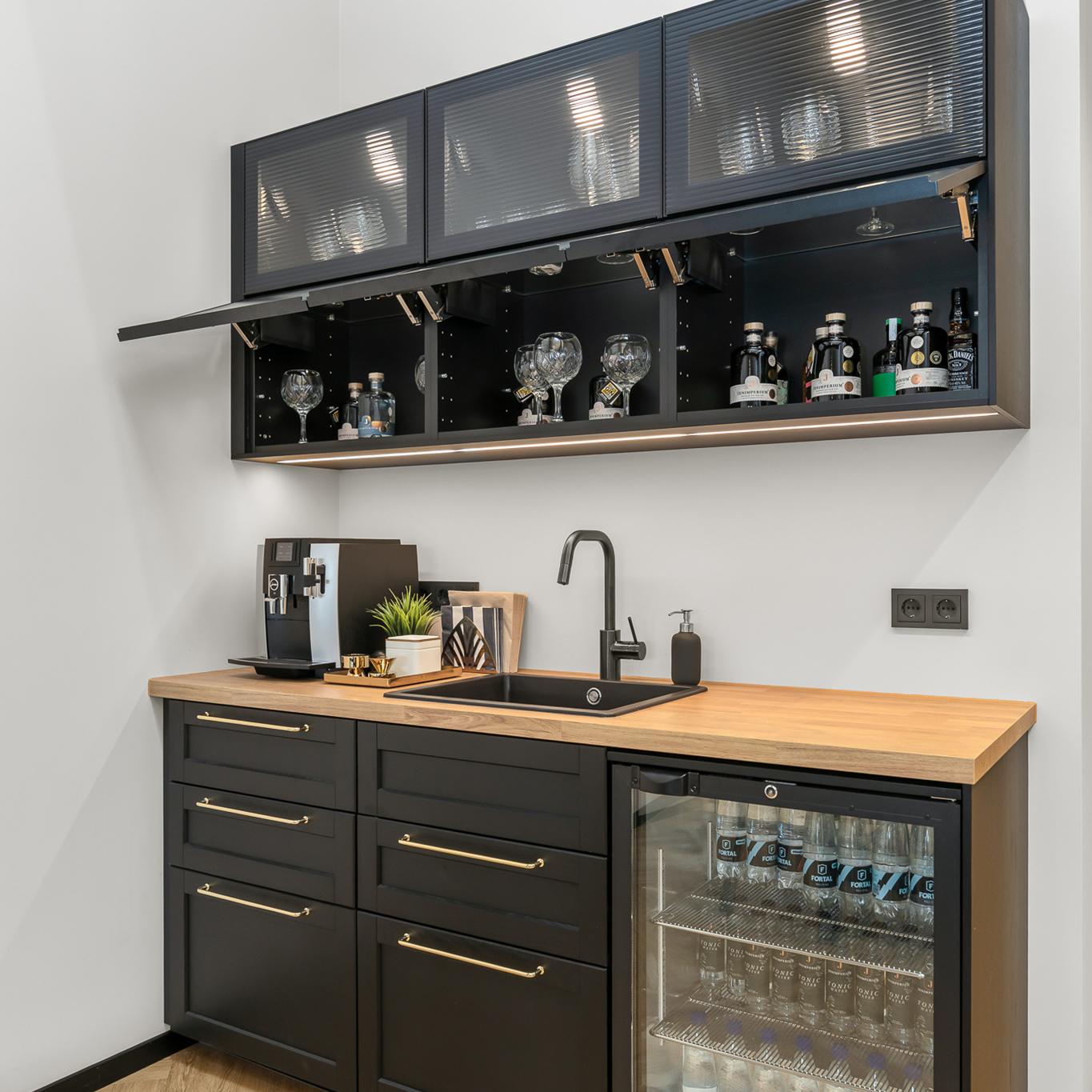 Compact kitchen with black cabinets and a wooden countertop, featuring a sink, coffee machine, and glass-front upper cabinets. A mini fridge stocked with drinks sits below, and glassware is visible inside the cabinets.