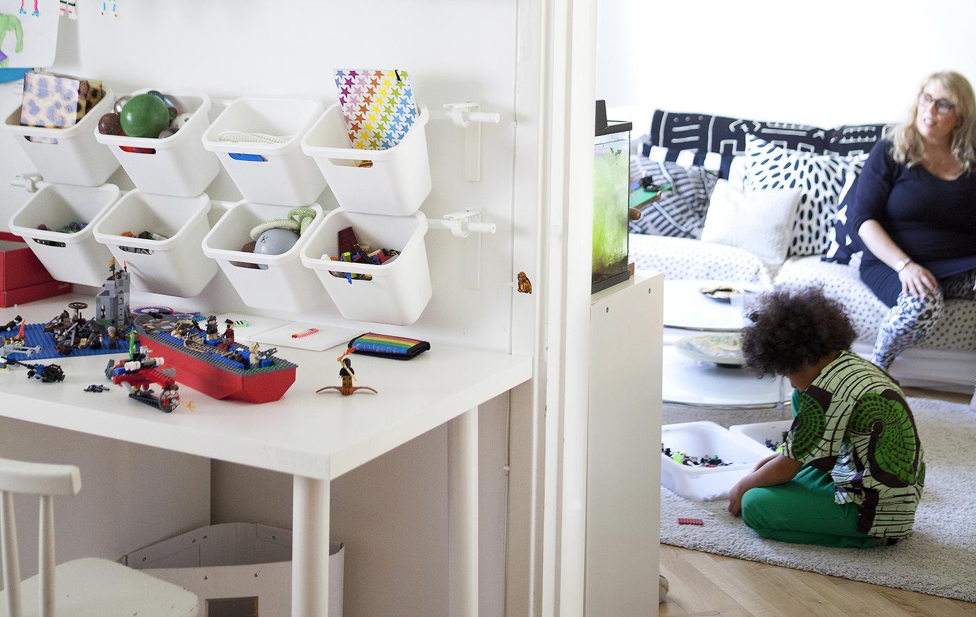 Containers hang on a wall-mounted rail to create toy storage in a child's bedroom. Mom and son sit in the background.