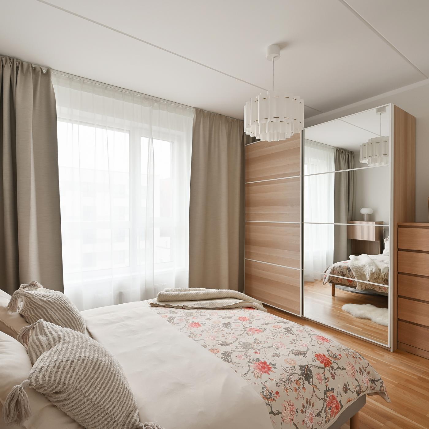 Contemporary bedroom with soft neutral tones, floral bedding, warm wooden furniture, mirrored wardrobe doors, and large windows that fill the space with natural light and create a calm, inviting atmosphere.