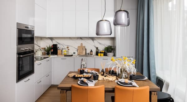 Contemporary kitchen and dining area with marble backsplash, wooden table, yellow floral centerpiece, and orange chairs.
