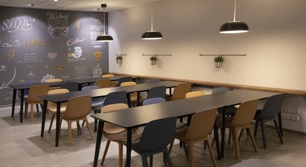 Contemporary office dining area with black tables, mixed-color chairs, and decorative wall art featuring food and coffee illustrations.