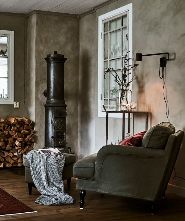 Corner of a grey-walled room with a green armchair, red cushion and footstool next to wood-burning stove and wood pile.