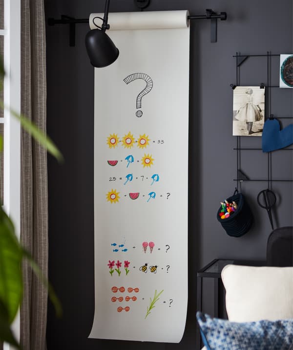 Corner of a room with a wall-mounted drawing-paper roll, a length of paper out with colourful, graphic maths problems.