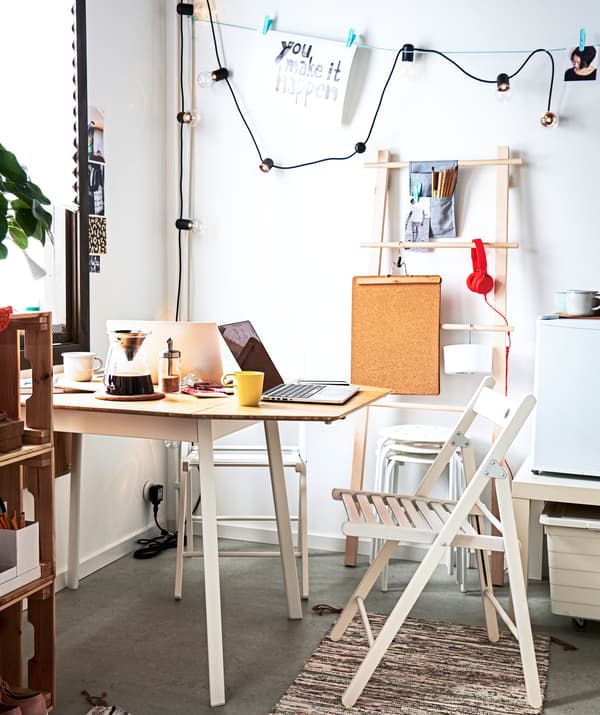 Corner of a room with table, one end to the wall, surrounded by folding chairs, extra stools and various storage.