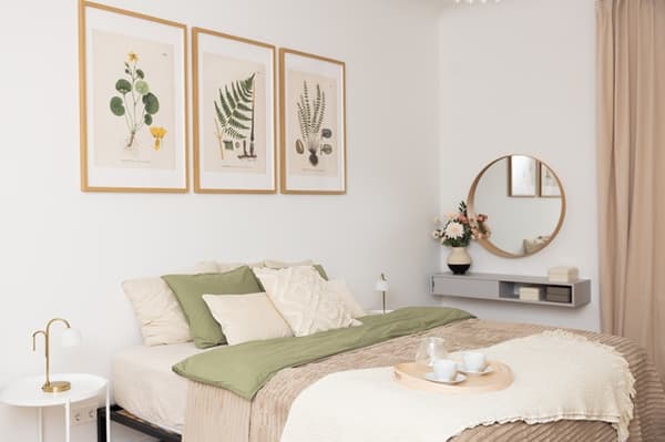 Cosy bedroom with a double bed, light textiles, decorative cushions, wall art and a round mirror above a table.