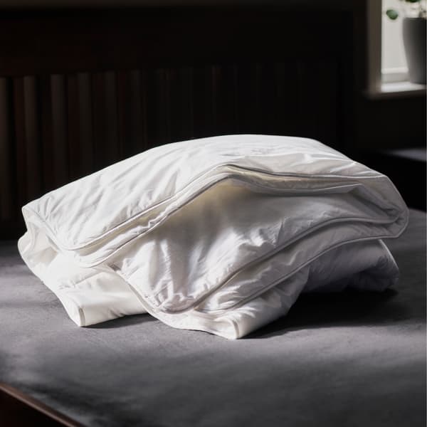Couette chaude STJÄRNBRÄCKA pliée sur un matelas gris, dans une pièce sombre. Une plante en pot est posée sur l’appui de fenêtre en arrière-plan.