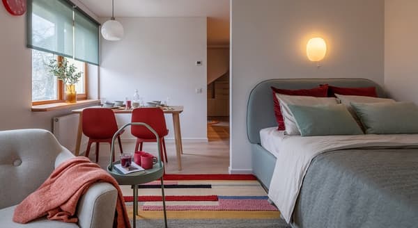 Cozy and colorful open-plan studio featuring a bedroom, dining area, and reading corner in a Scandinavian style. On the right, a soft gray upholstered bed is decorated with sage green and burgundy pillows, beneath a warm wall-mounted light. A striped multicolored rug unifies the space, stretching toward a light gray armchair with a coral throw and a small green round side table holding mugs and a book. The dining area in the back includes a wooden table set for four, with red chairs and a large amber vase with greenery on the window sill. Soft daylight filters through green roller blinds, creating a calm, inviting atmosphere.