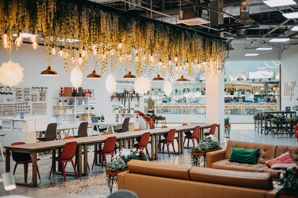 Cozy and modern IKEA lounge area with long communal wooden tables, red chairs, hanging greenery, and warm pendant lighting.