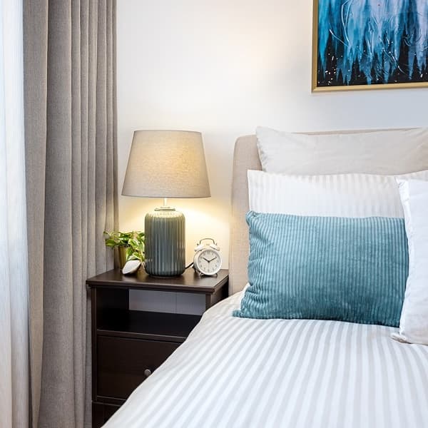Cozy bedroom corner with striped bedding, teal pillow, bedside lamp, and alarm clock on dark wood nightstand.