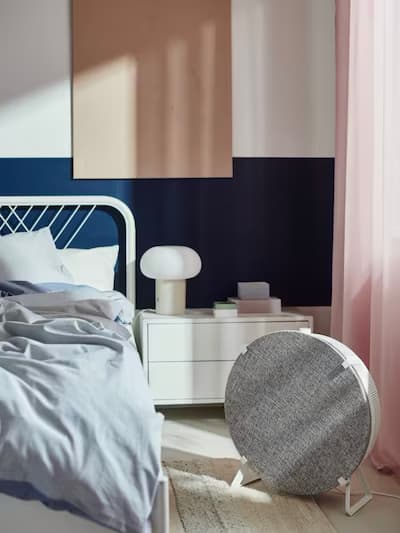 Cozy bedroom with white metal bed frame, light blue bedding, white nightstand with lamp and books, navy and white walls, pink curtains, and a round gray speaker.