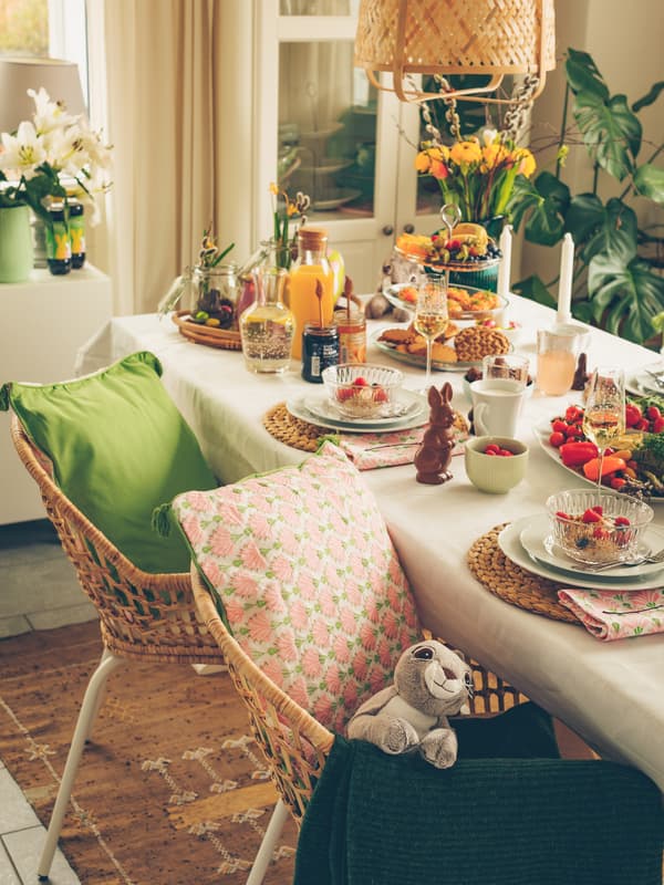 Cozy dining table set with colorful cushions, breakfast dishes, fresh flowers, and decorative items in a sunlit room.