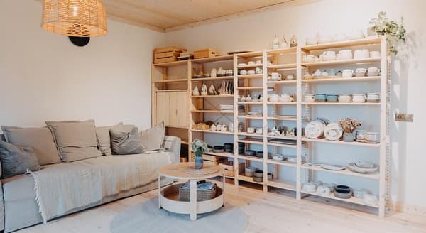 Cozy living room with beige sofa, wooden shelves full of handmade ceramic tableware, and warm natural lighting.