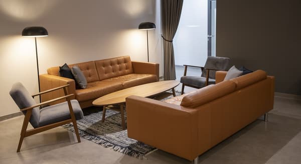 Cozy office lounge with two brown leather sofas, grey armchairs, oval wooden table, and floor lamps.