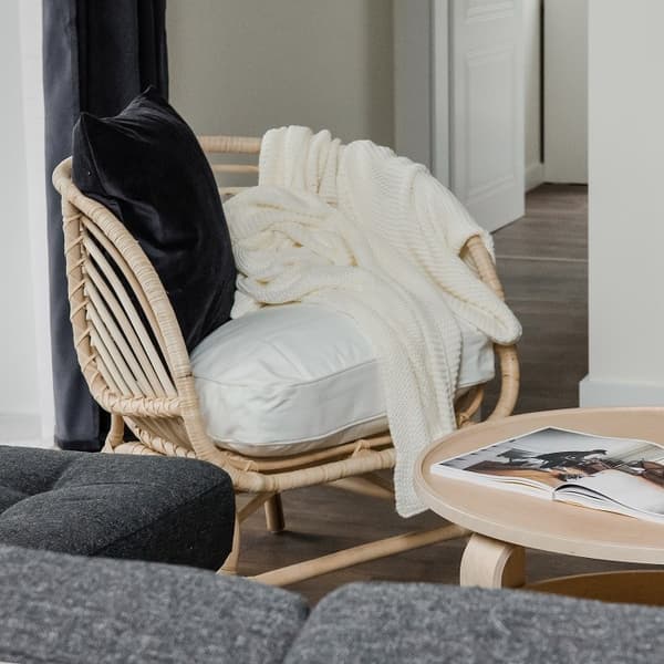 Cozy reading nook featuring a natural rattan armchair with white cushions, a dark velvet pillow, and a chunky cream knit throw draped over the back. Next to it is a light wooden round coffee table with an open magazine on top. The edge of a dark gray sofa is visible in the foreground, and dark navy curtains hang to the left. The setting exudes warmth and relaxation in a modern, Scandinavian-style interior.