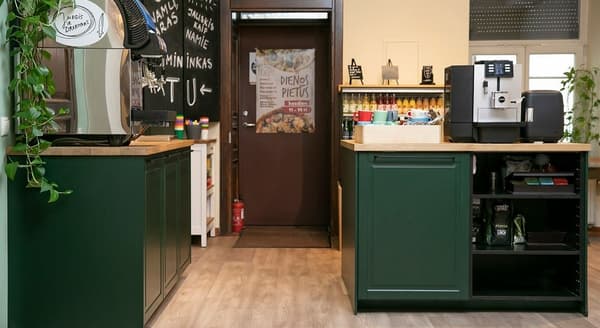 Cozy self-service coffee station with green cabinetry, espresso and vending machines, colorful mugs, wooden countertops, beverage fridge, chalkboard signage, and indoor plants on wood flooring.