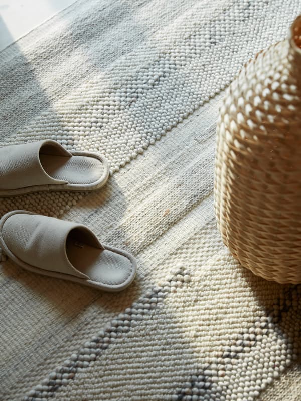 Cream-coloured slippers and a woven bamboo vase stand on a hand-woven BRÖNDEN rug.