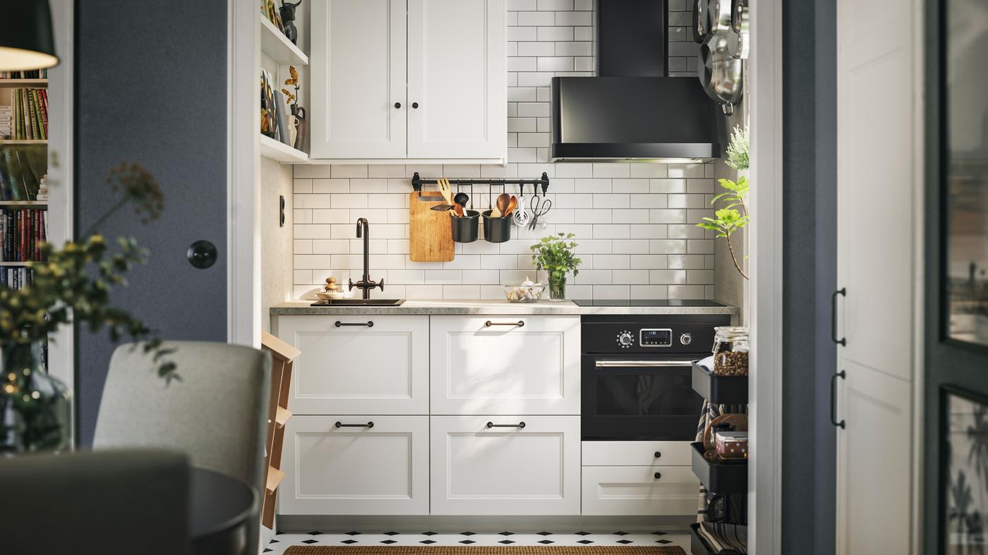 Cuisine carrelée blanche avec façades AXSTAD blanc mat, armoires hautes, électroménagers noirs et étagères murales.