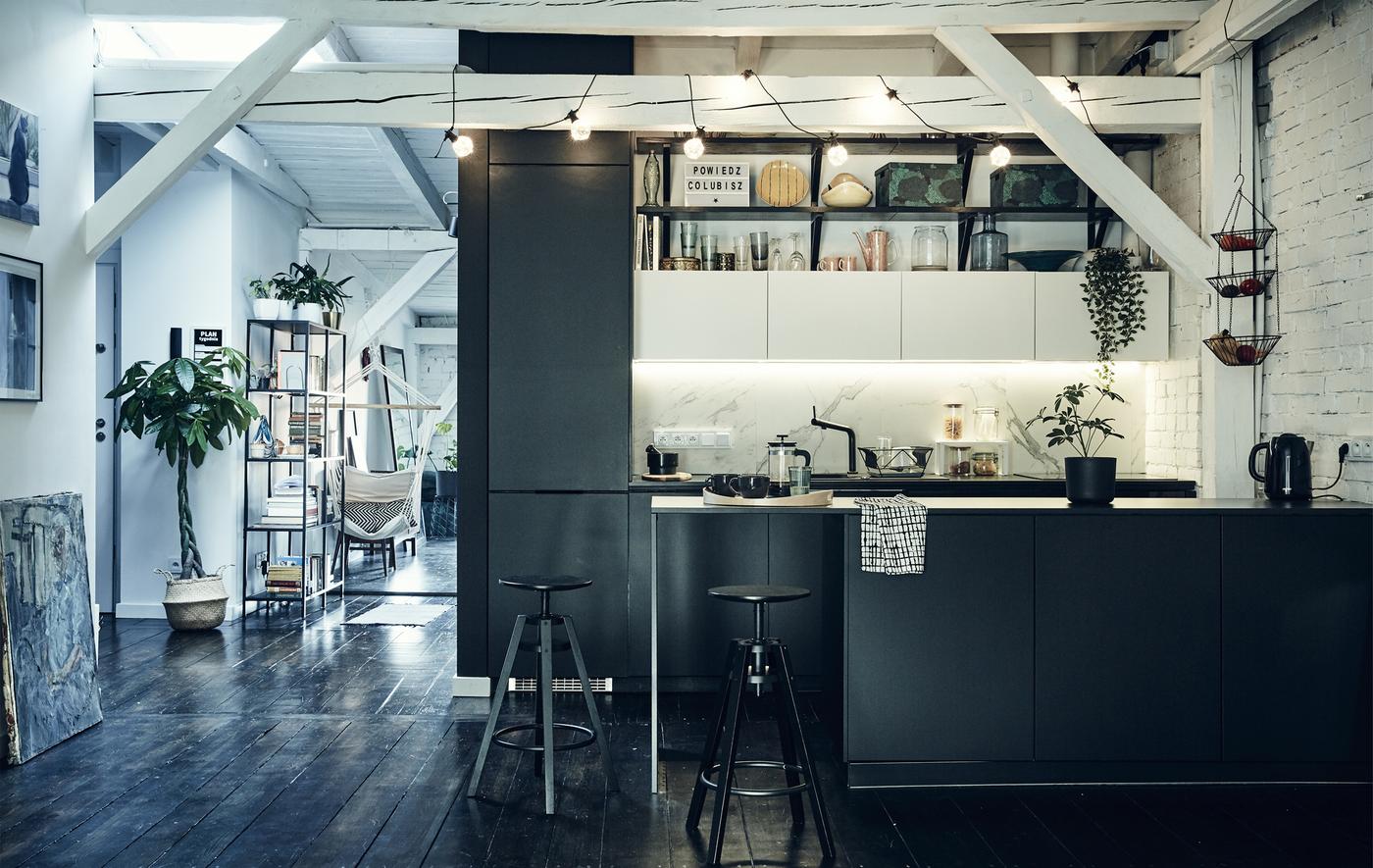 Cuisine noire dans un espace décloisonné, avec poutres apparentes blanches et sols en bois foncé.