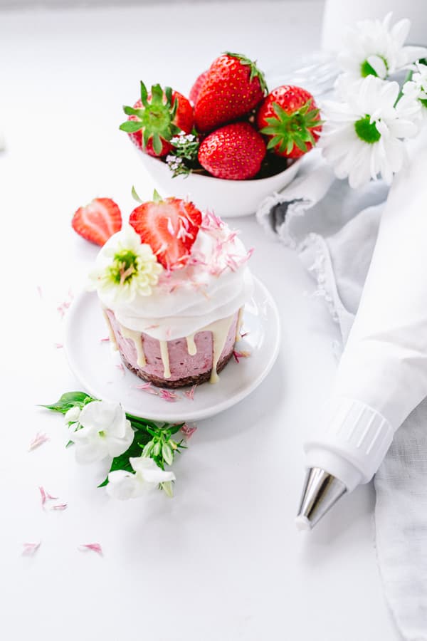 Cupcake with cream and strawberry on white plate, bowl of strawberries, flowers and piping bag in background.