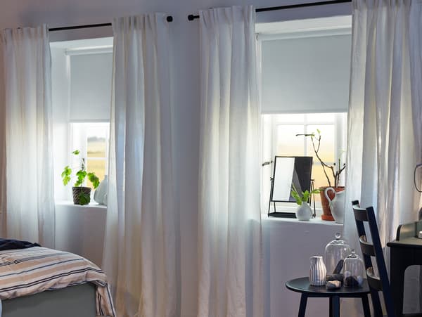 Curtains in white are hanging on black curtain rods in two bedroom windows where also white roller blinds are mounted.