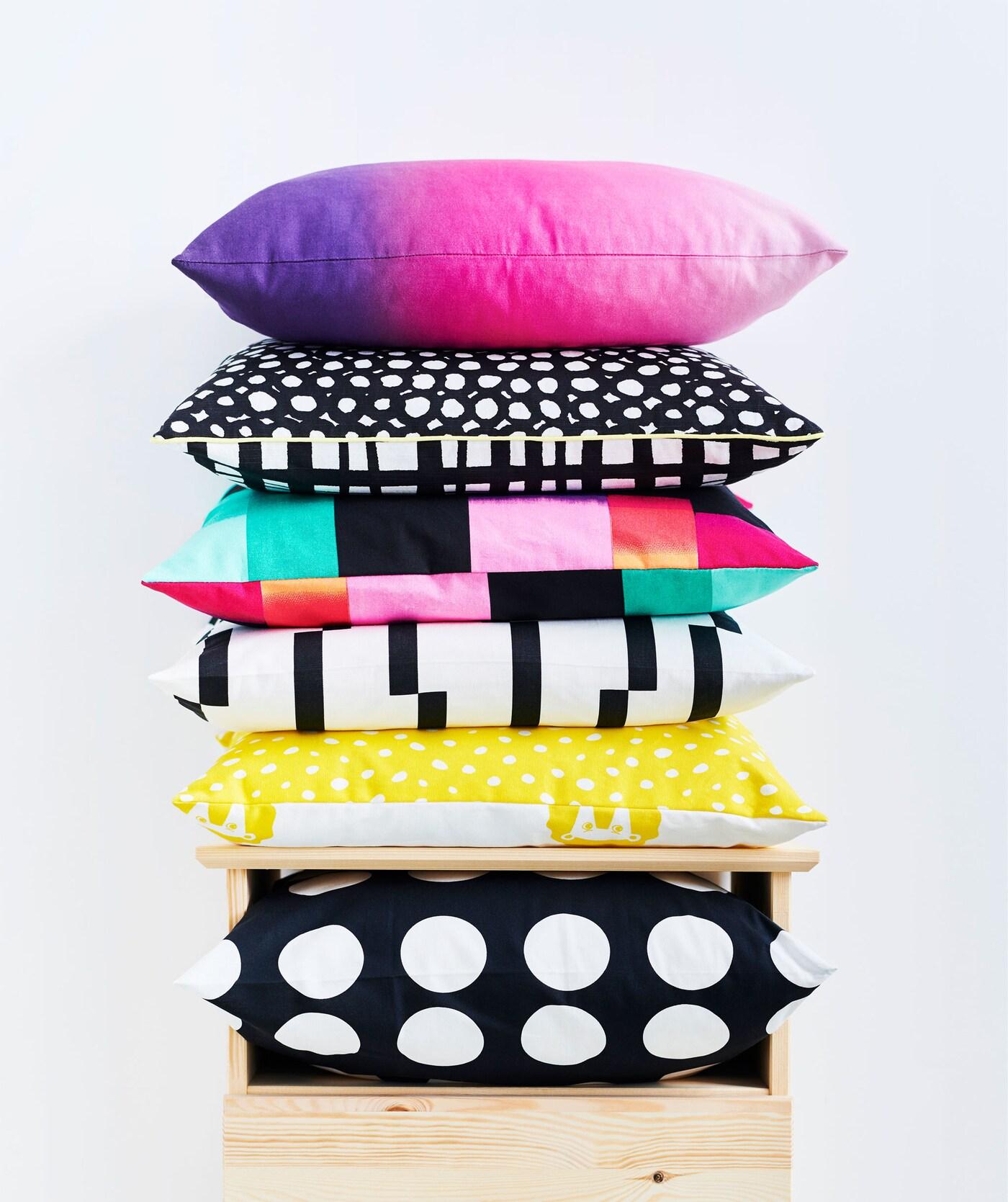 Cushions – black and white, graphic patterns, sharp coloring in a mix – piled in a stack on top of  a chest of drawers.