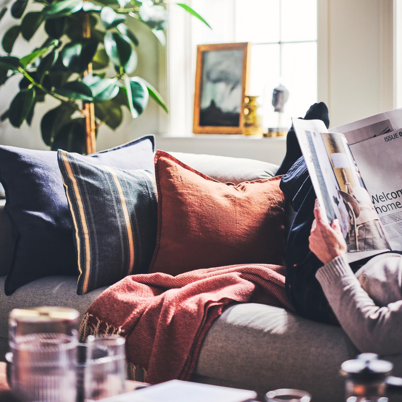 Cushions dressed in a STORTIMJAN cushion cover and DYTÅG cushion covers placed on a sofa next to a person reading a magazine.