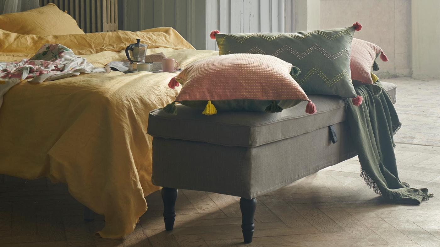 Cushions in KLARAFINA and MOAKAJSA covers lie on a STOCKSUND bench at the foot of a bed with yellow PUDERVIVA bed linen.