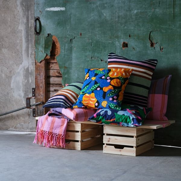 Cushions with MÄVINN covers in different patterns are placed on wooden boxes against a weathered green plaster wall.