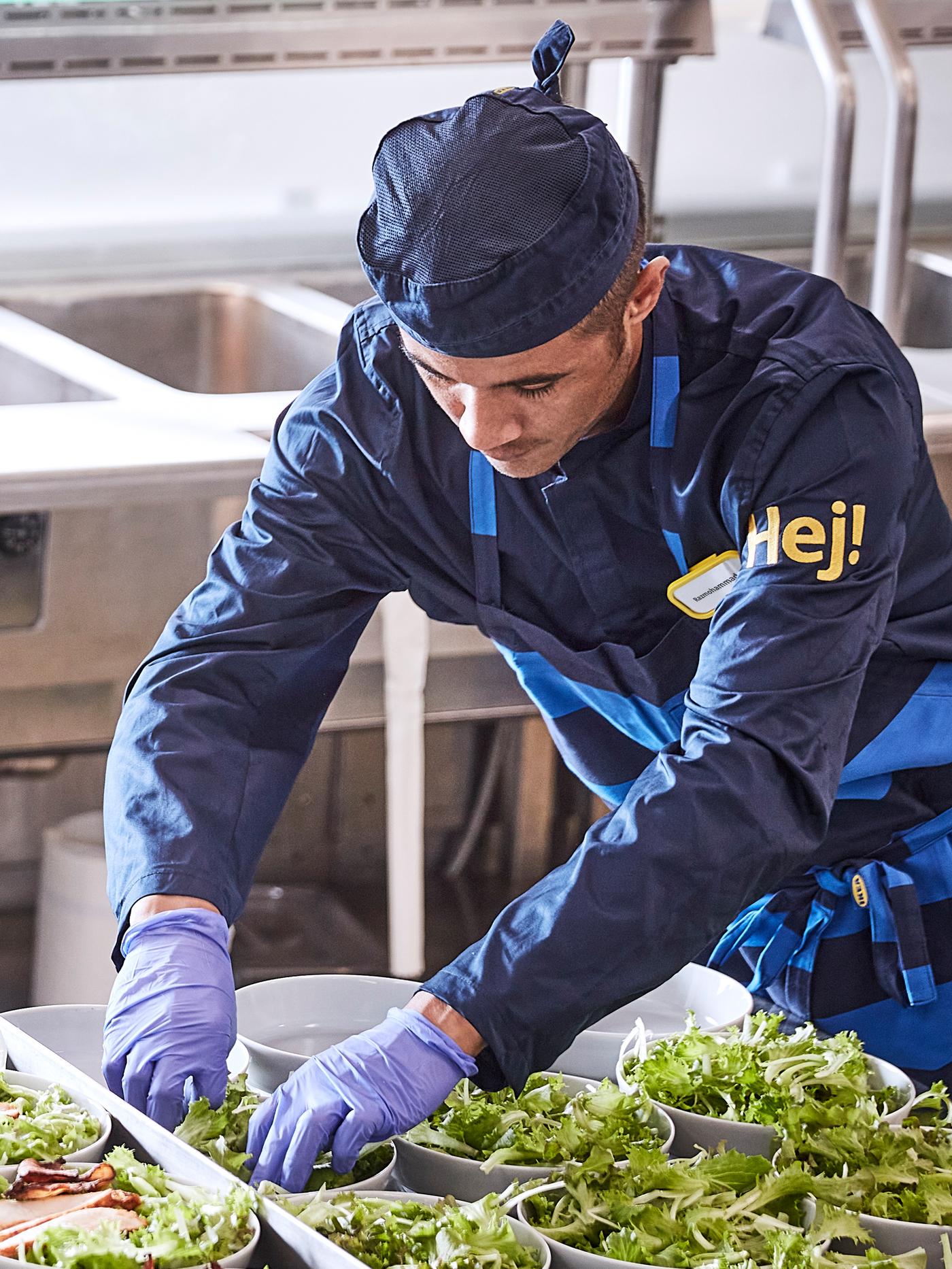 Dans la cuisine d’un restaurant, une personne vêtue d’une tenue de travail, de gants et d’un tablier remplit des bols de salade.