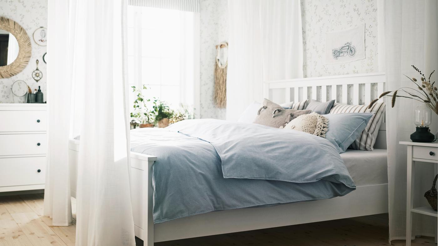 Dans une chambre à coucher claire, un lit HEMNES blanc entouré de voilages SILVERLÖNN blancs et garni de coussins et de linge lit bleu.