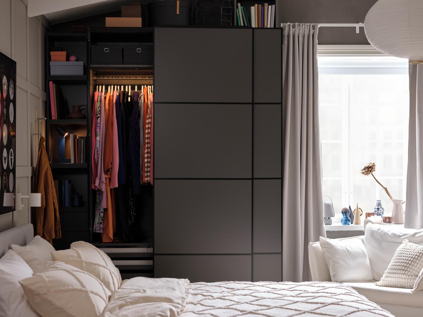 Dans une chambre de couleur grise, une armoire PAX/MEHAMN gris foncé avec une porte ouverte. L’éclairage intérieur éclaire des vêtements suspendus à une tringle.