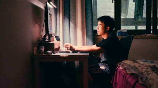 Dans une pièce sombre d’un appartement urbain, jeune homme assis à un bureau avec un clavier, regardant vers un écran d’ordinateur.
