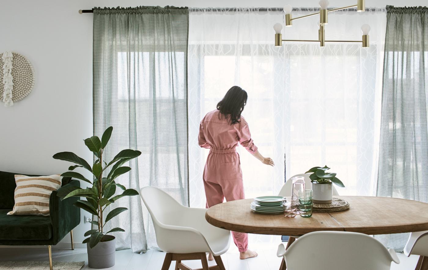 Dans une salle à manger avec une table en bois et des chaises blanches, une femme vêtue d’une combinaison rose arrange les rideaux de la porte-fenêtre. 