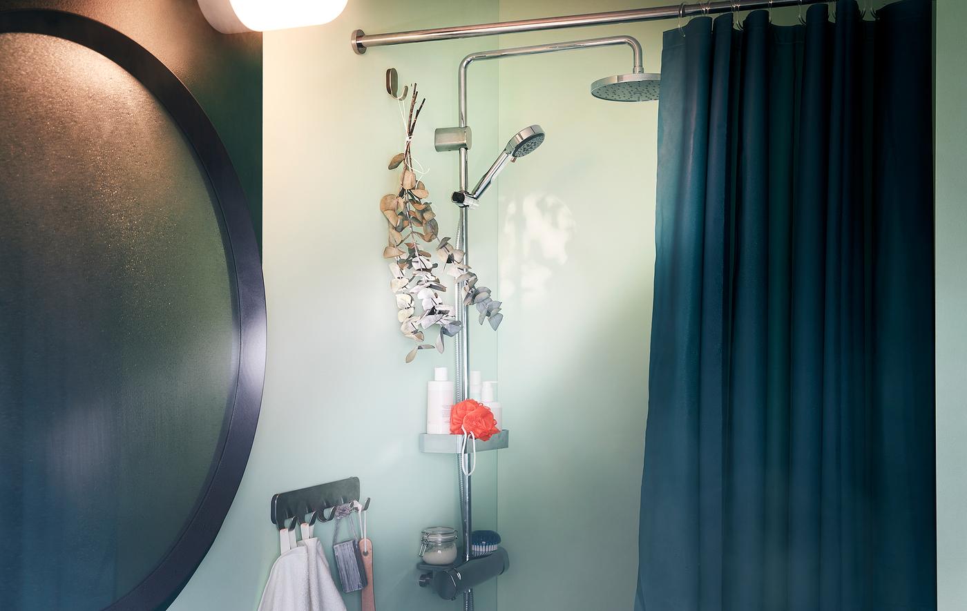 Dans une salle de bains, un miroir embué à côté d’une douche avec des accessoires ; un bouquet de branches d’eucalyptus est suspendu sur un crochet.