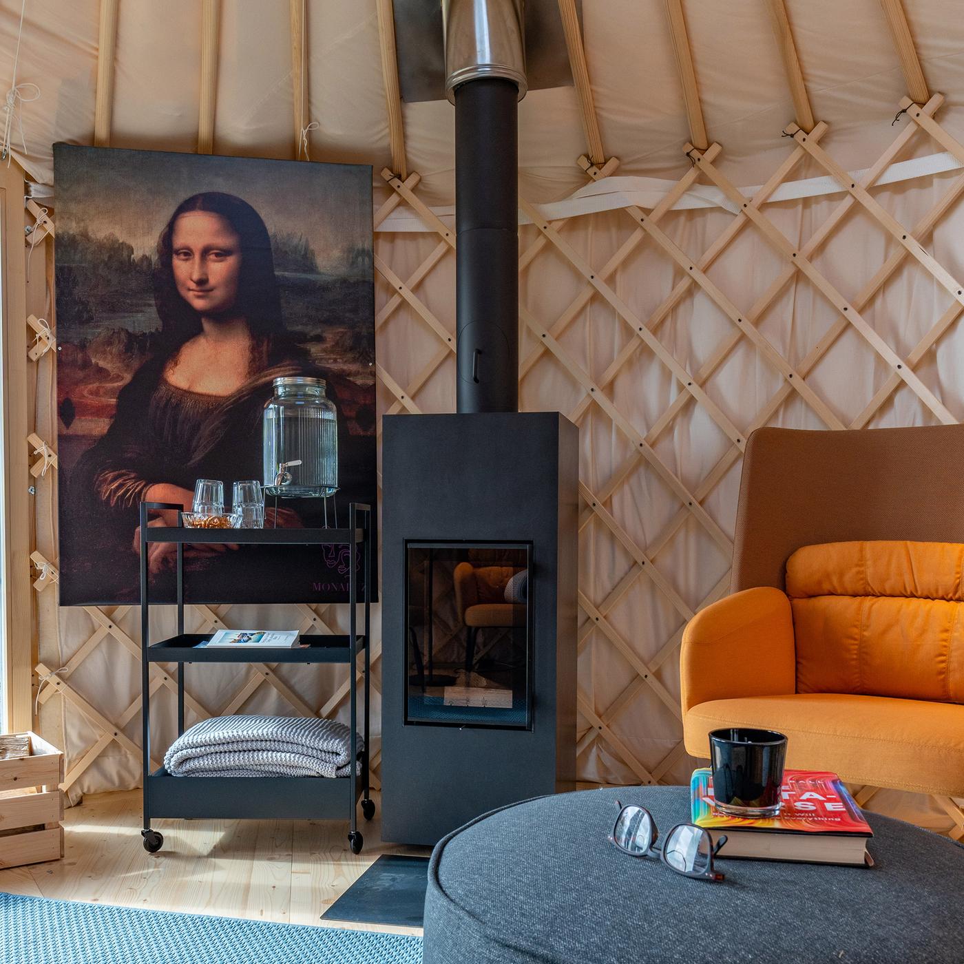 Dans une yourte se trouve un poêle à bois à côté d’un grand tableau de la Joconde ainsi qu’un fauteuil avec une table d’appoint