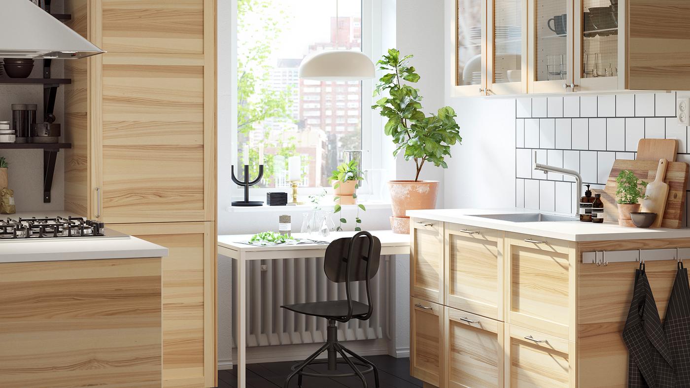 Dapur kecil yang terang dengan kabinet kaca dan kayu ash semula jadi yang tradisional dan meja berwarna putih di hadapan tingkap.
