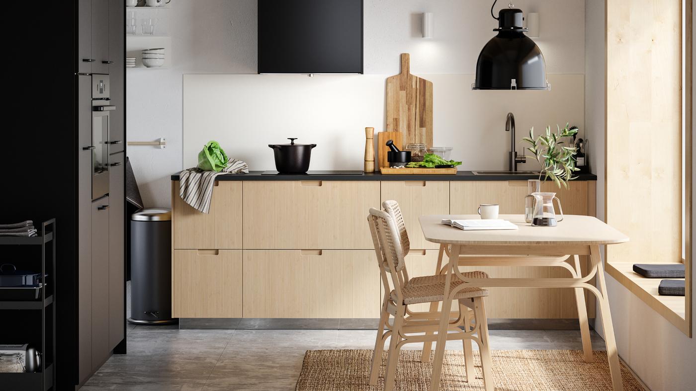 Dapur METOD dengan bahagian depan FRÖJERED yang dibuat daripada buluh berwarna cerah, meja dan dua kerusi serta lampu pendan SVARTNORA berwarna hitam.