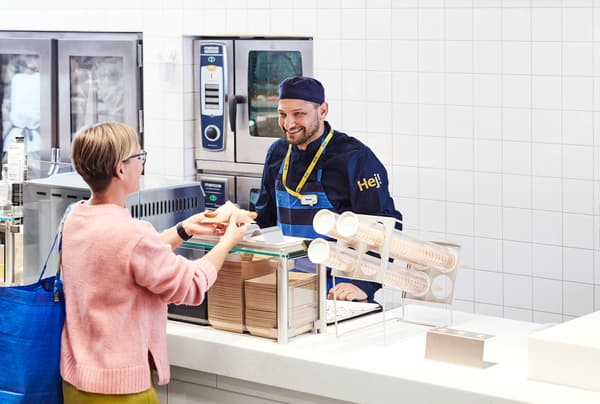 Das Bild zeigt eine freundliche Szene an der IKEA Bistro-Theke: Eine Kundin mit blauer IKEA-Tasche erhält ihr Essen von einem lächelnden Mitarbeiter in IKEA-Uniform.