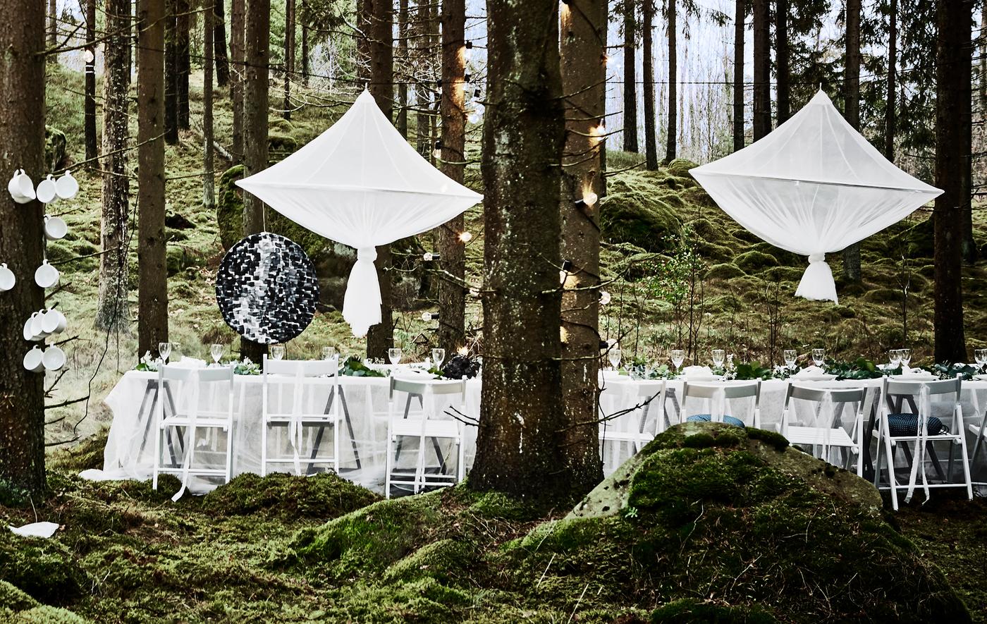 Décor de table pour une fête de mariage en forêt, avec chaises blanches, nappes en voilages et grandes décorations centrales suspendues à la manière de lustres