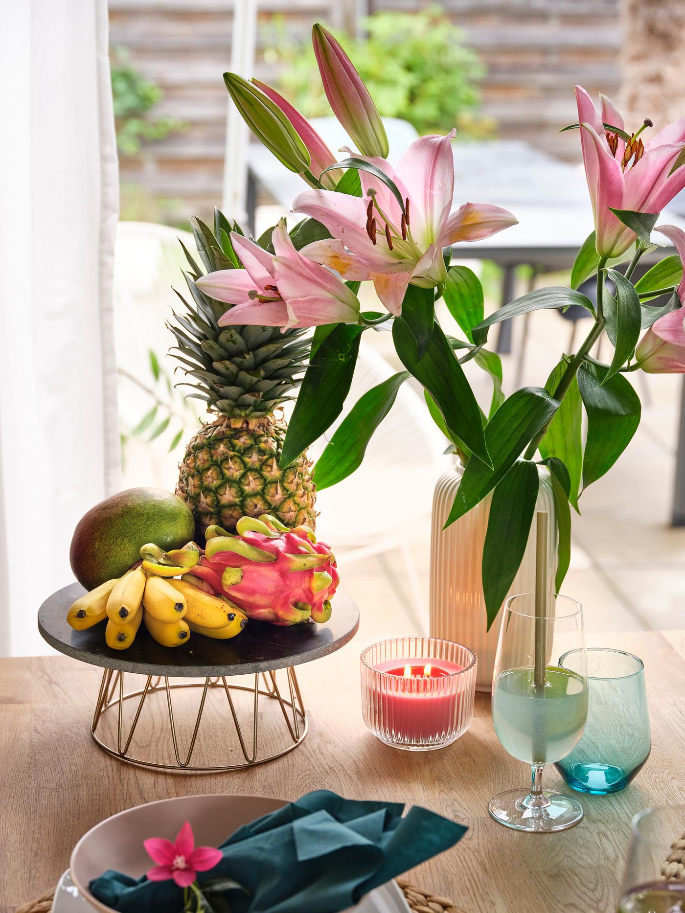 Décoration de table tropicale avec des lys roses, un ananas, un fruit du dragon, des bananes, une bougie et des verres colorés.