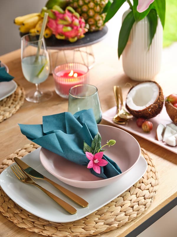 Décoration de table tropicale avec un bol rose, une serviette turquoise, des couverts dorés, une noix de coco et une bougie.