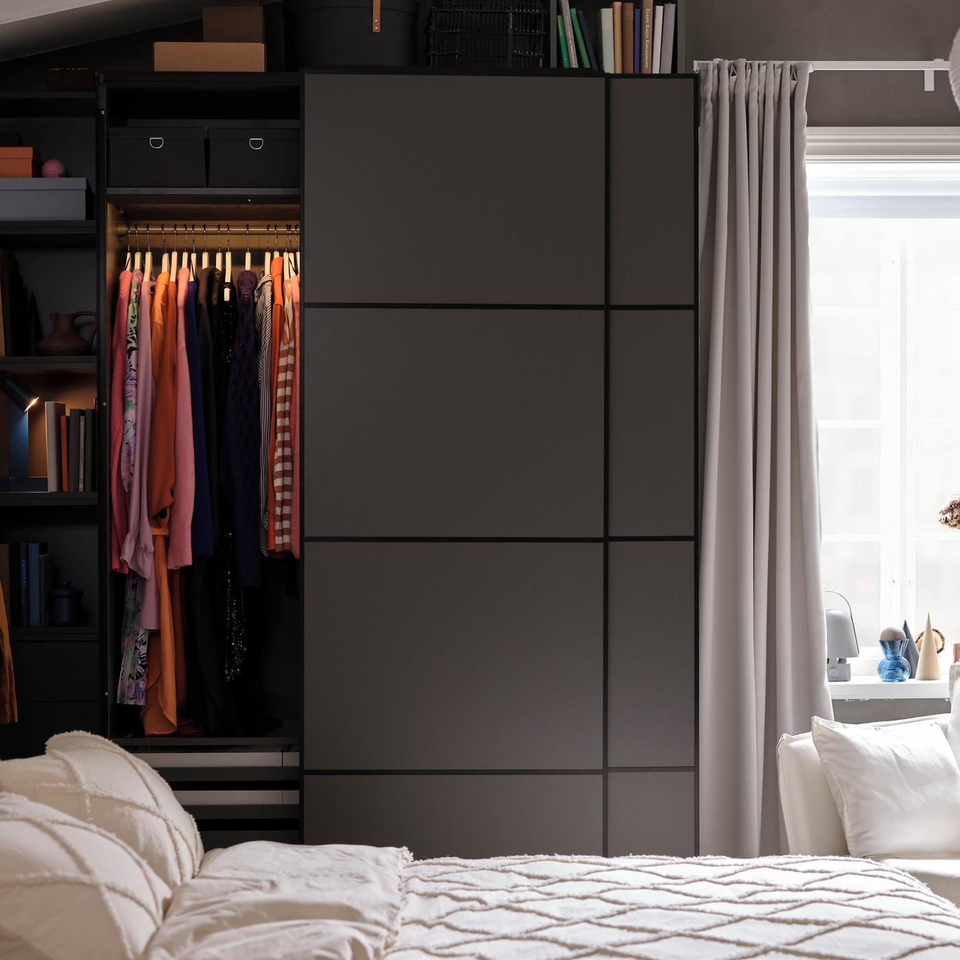 Derrière un lit, une armoire-penderie PAX/MEHAMN gris foncé dont la porte coulissante gauche ouverte révèle des vêtements suspendus à une tringle.