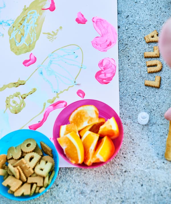 Des bols avec des biscuits en forme de lettres et des quartiers d'orange, et une peinture d'enfant, à côté de biscuits épelant LULA sur un sol en béton.