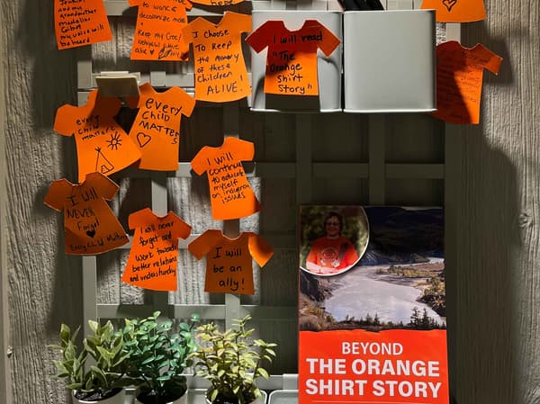 Des bouts de papier orange en forme de chandail sont disposés sur un mur d’exposition. Il y a un mot écrit sur chacun. 
