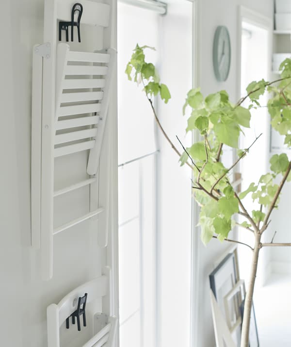 Des chaises pliantes blanches suspendues à des crochets en forme de mini chaises sur un mur blanc à côté d'une plante d'intérieur.