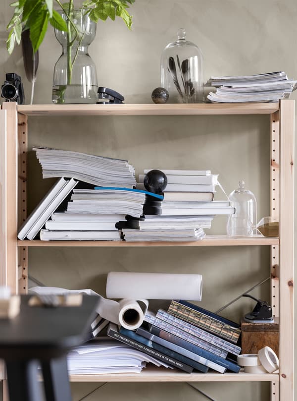 Des étagères en bois montées au mur, sur lesquelles se trouvent des piles désordonnées de livres et de magazines, ainsi que des vases et cloches en verre avec des fleurs et des plumes.